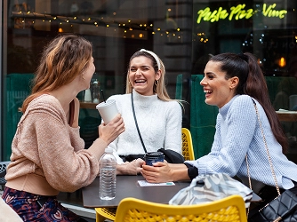 Three women sit at an outdoor table, laughing and holding coffee cups